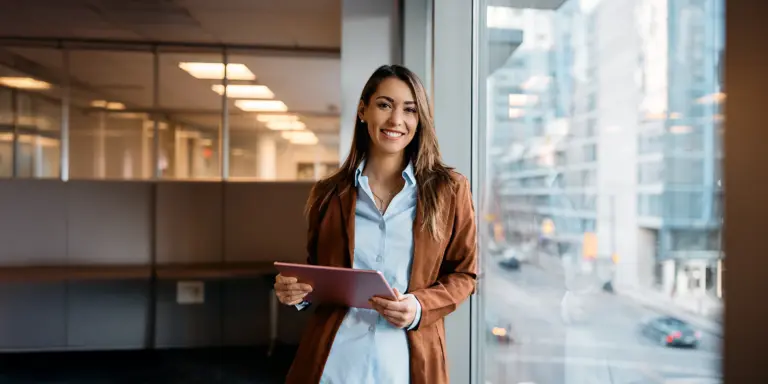 Eine lächelnde Frau im Büro hält ein Tablet, steht am Fenster mit Stadtblick im Hintergrund.