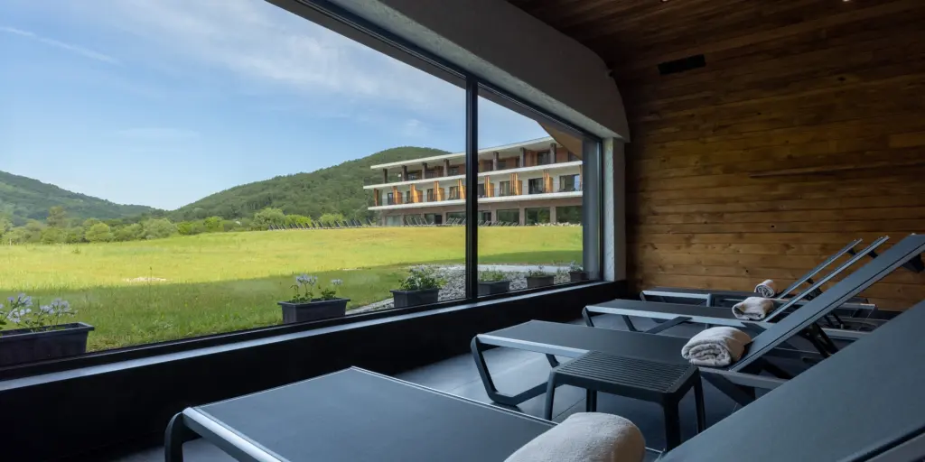 Entspannungsbereich mit Liegen, Blick auf Wiesen und modernes Gebäude, umgeben von grünen Hügeln.